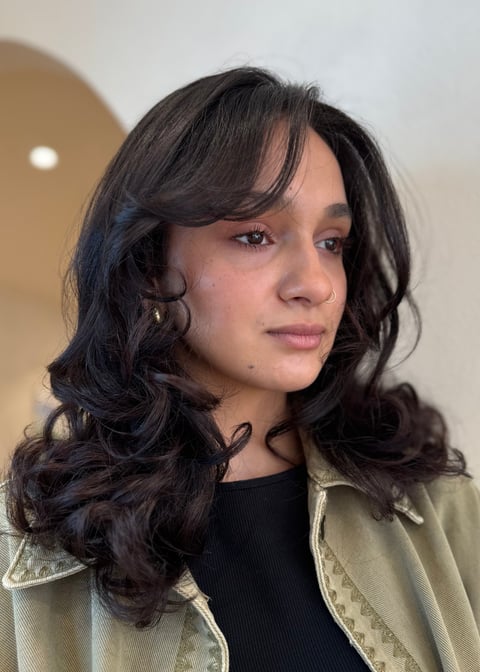 Portrait of a young woman with long wavy black hair, wearing a black top and beige jacket, looking to the side