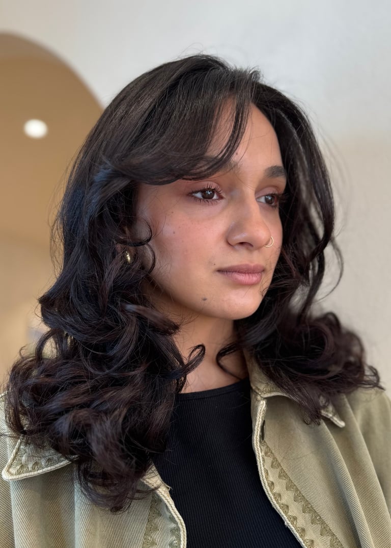Portrait of a young woman with long wavy black hair, wearing a black top and beige jacket, looking to the side