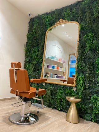 Modern salon interior with orange styling chair, large wall-mounted mirror, green plant backdrop, and gold accent table