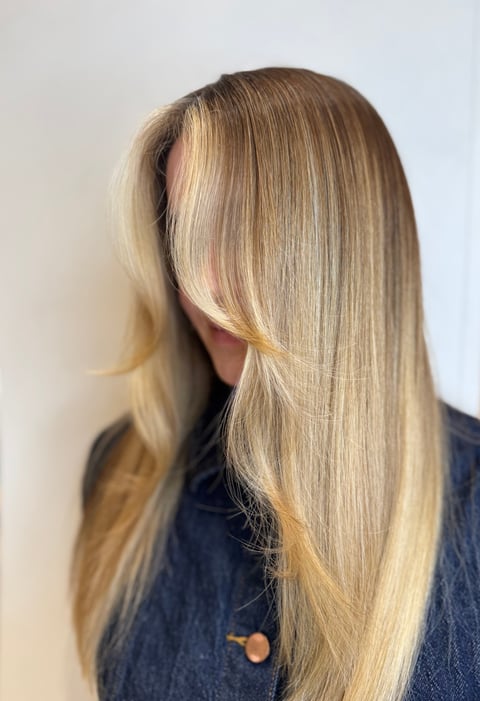 Person with long blonde straight hair covering face, wearing dark blue top, photographed from side profile against light background