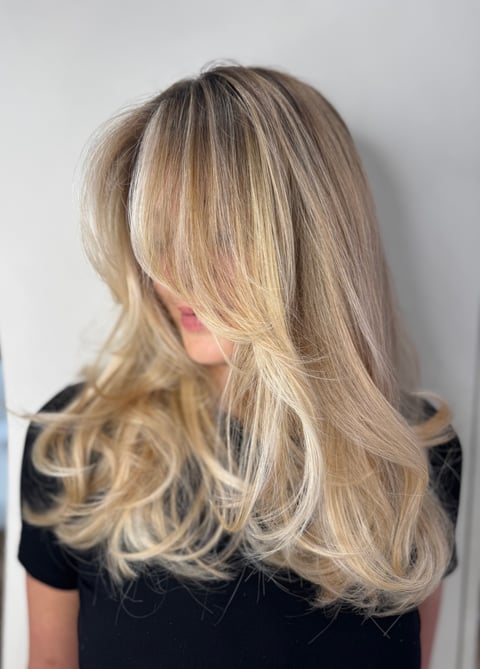 Person with long blonde layered hair with highlights, wearing a black shirt, photographed from behind against a white background